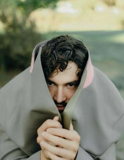 Close-up of a man with dark hair, light skin, and a mustache, partially covered by a grey hooded cape in an outdoor setting.
