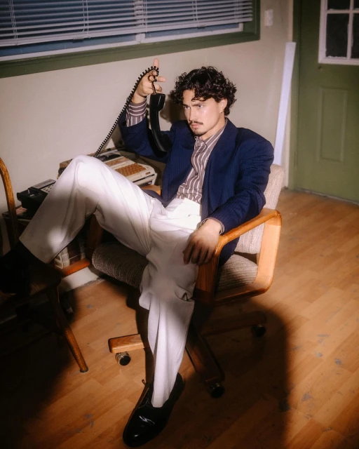 Stylish man in blazer sits in office chair holding landline phone, with striped shirt, white pants, and dark loafers.