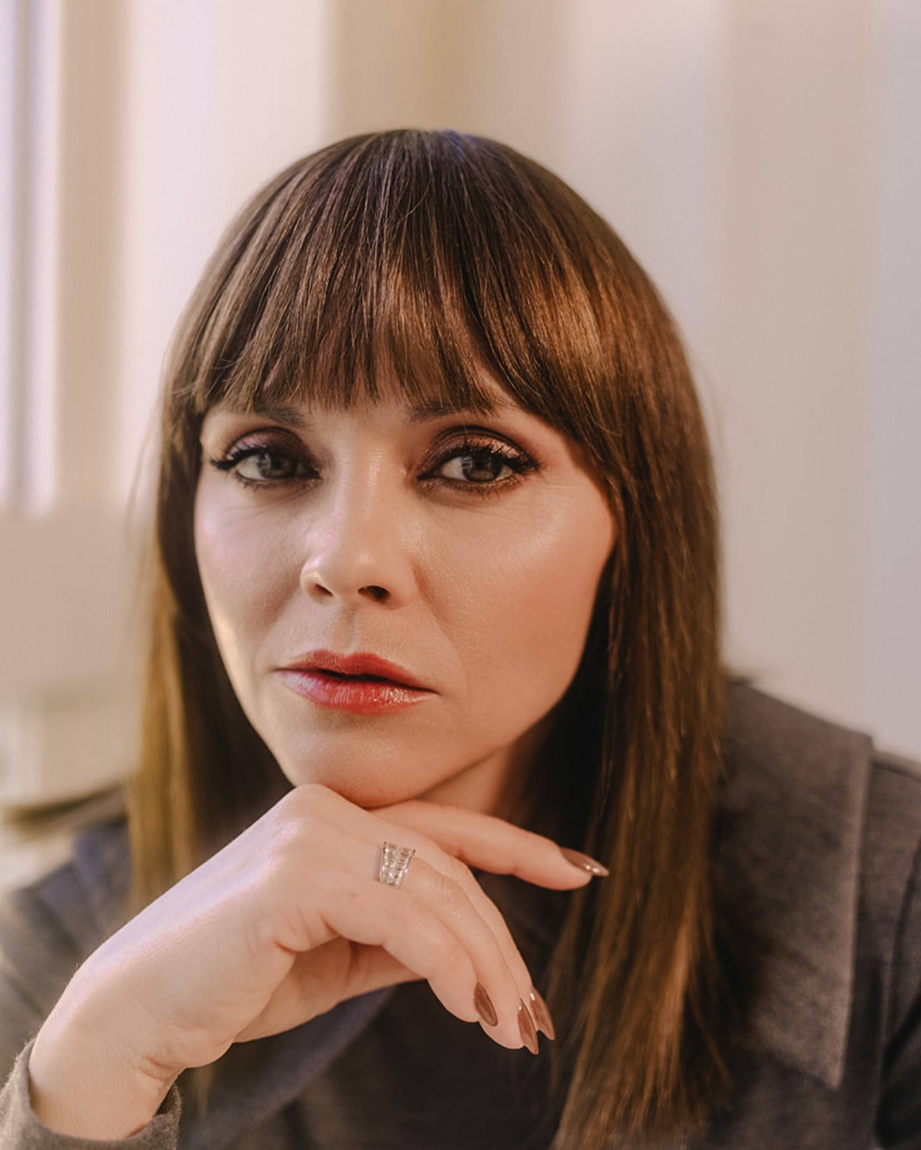 Close-up portrait of a woman with brown hair and bangs, resting her chin on her hand, wearing a ring on her finger.