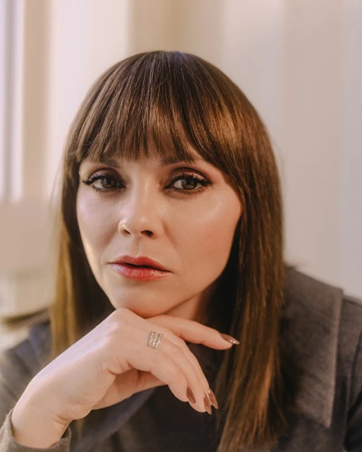 Close-up portrait of a woman with brown hair and bangs, resting her chin on her hand, wearing a ring on her finger.
