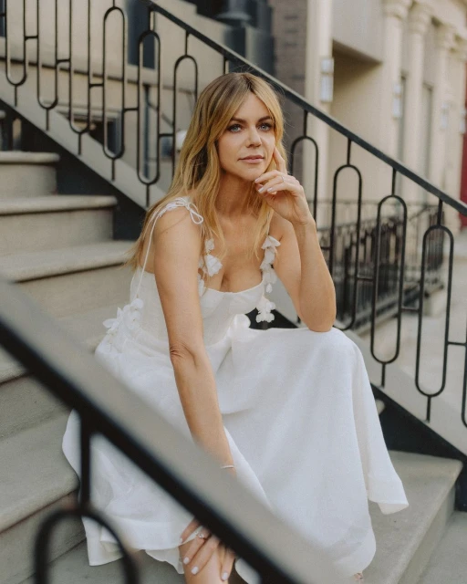 A fair-skinned woman with blonde hair sits on outdoor steps wearing a white dress with floral details, with a city street backdrop.