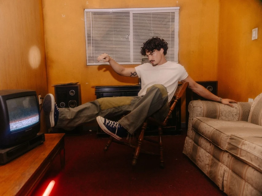 An adult male with curly hair sits backward in a chair indoors, wearing casual clothing, with a vintage TV and furniture in the background.