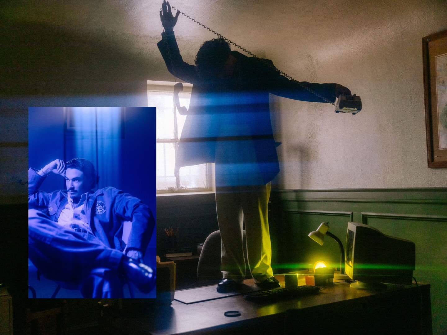 Man in suit standing on desk and talking on phone; framed portrait of seated man, blue lighting, office setting, interior.