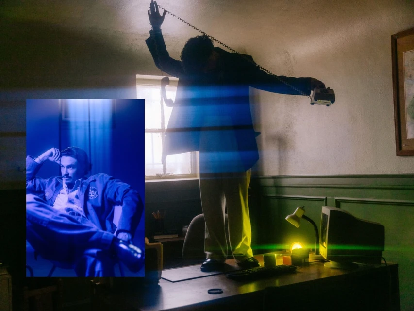 Man in suit standing on desk and talking on phone; framed portrait of seated man, blue lighting, office setting, interior.
