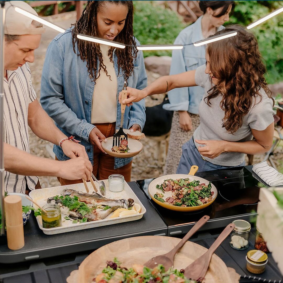 A group of four people prepare food outdoors on a portable cooking station, featuring trout, salad, and vegetables in natural light.