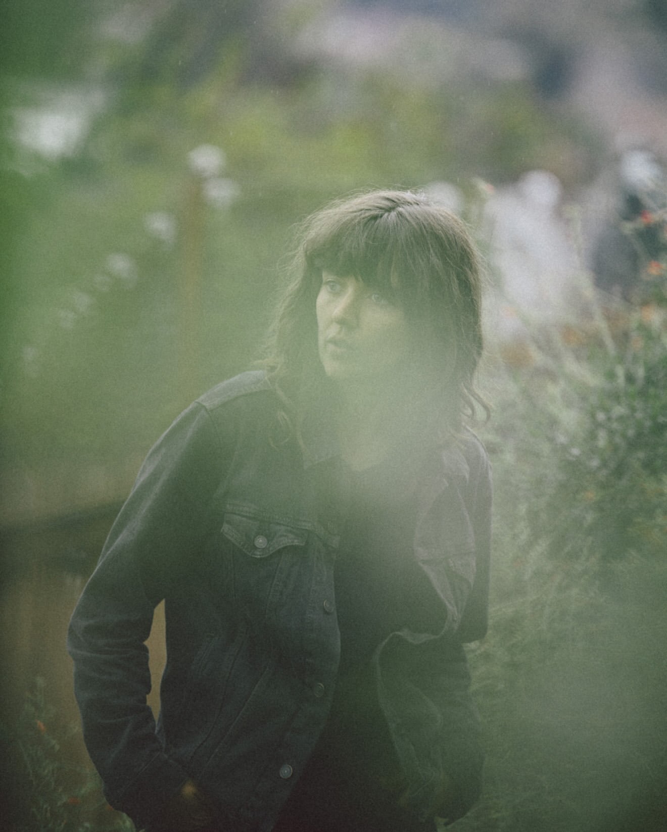 Adult female with bangs and denim jacket gazes away in a blurred outdoor setting with trees and foliage, creating a portrait-style image.