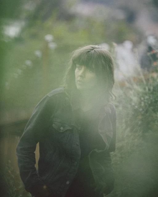 Adult female with bangs and denim jacket gazes away in a blurred outdoor setting with trees and foliage, creating a portrait-style image.