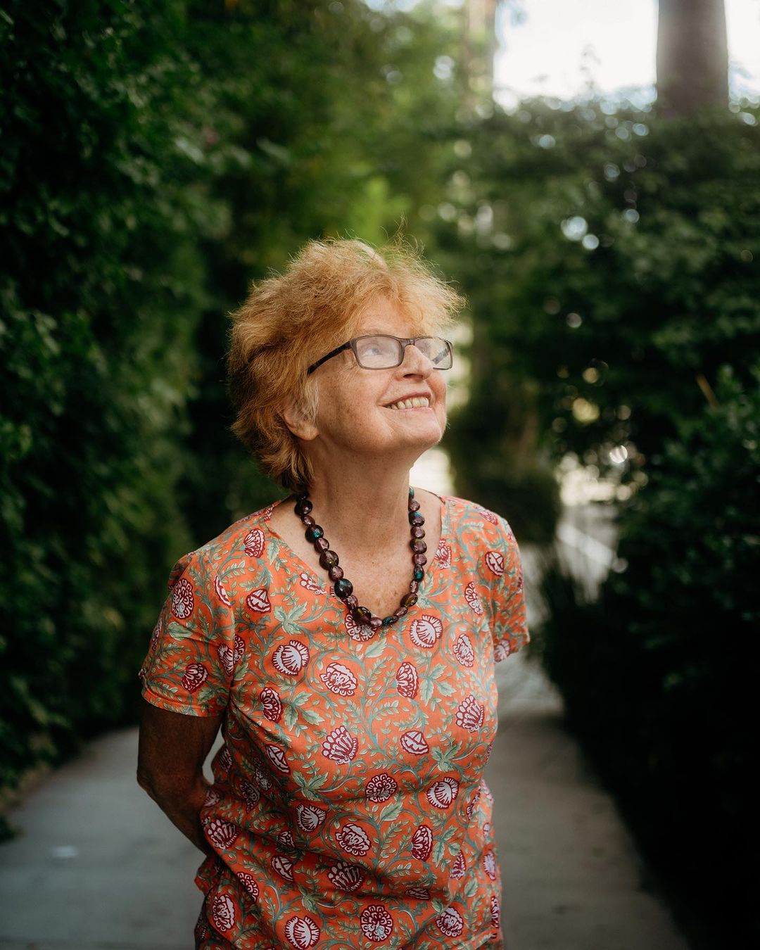 Elderly woman wearing eyeglasses and patterned orange top standing outdoors near lush green foliage.
