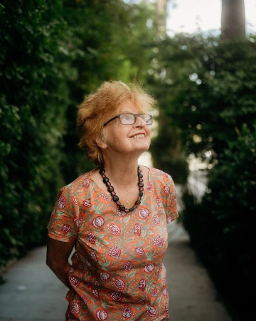 Elderly woman wearing eyeglasses and patterned orange top standing outdoors near lush green foliage.