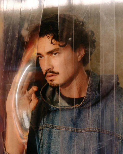 Portrait of a young adult male with curly hair and a moustache, wearing a denim jacket, standing indoors and looking off to the side.