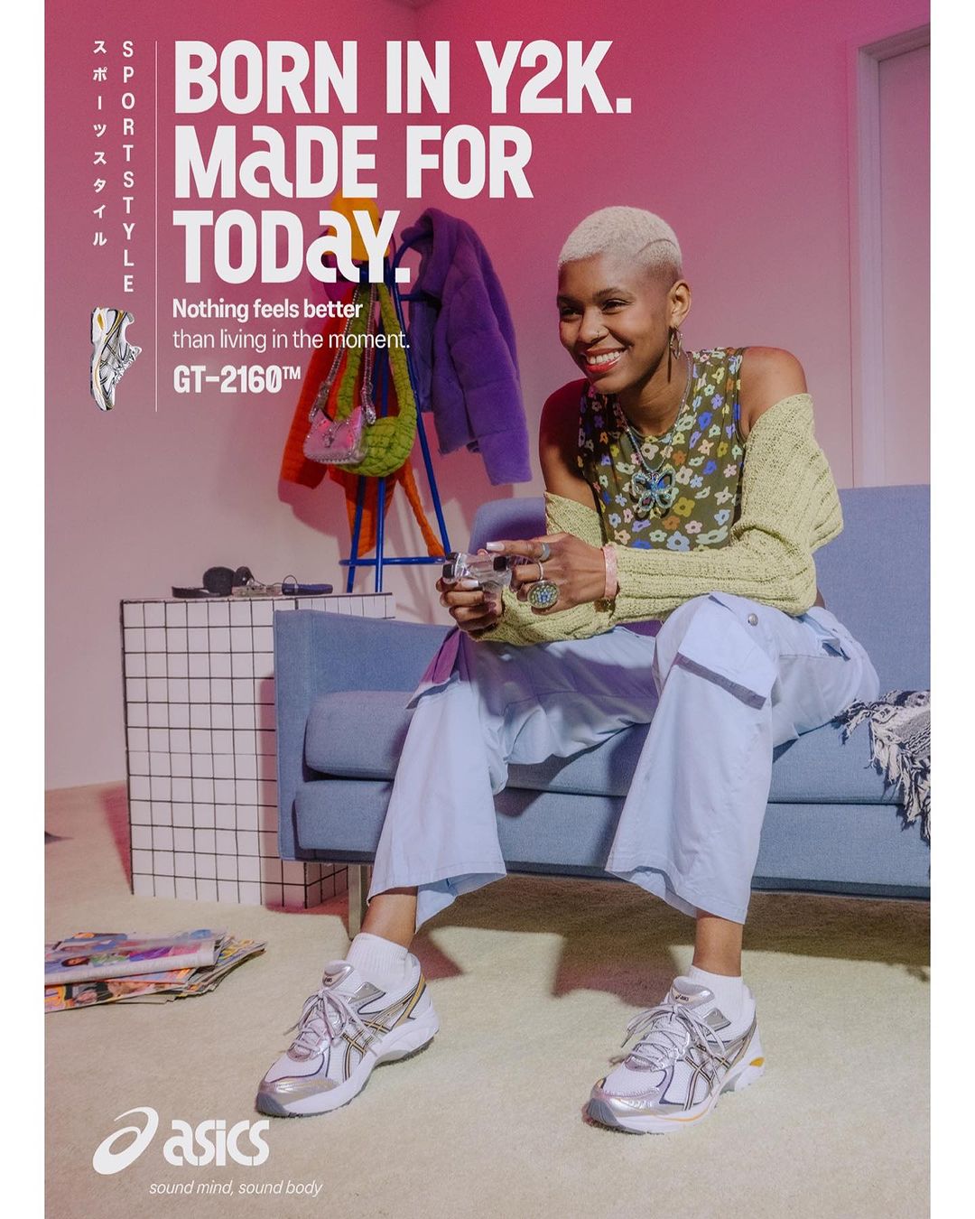 Woman in casual outfit sits on blue sofa next to silver Asics sneakers and graphic wall; indoor setting with pink wall and natural light.
