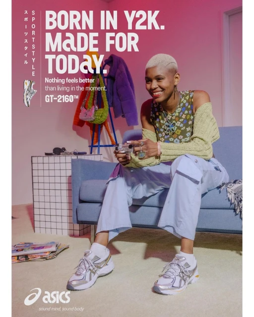 Woman in casual outfit sits on blue sofa next to silver Asics sneakers and graphic wall; indoor setting with pink wall and natural light.
