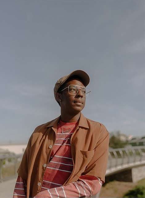Adult male in casual outdoor portrait wearing a brown shirt, hat, and glasses, looking off into the distance, with a bridge in background.