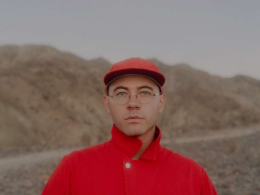 Portrait of a person wearing red hat and jacket outdoors against a backdrop of desert hills, showcasing simple clothing and a neutral expression.