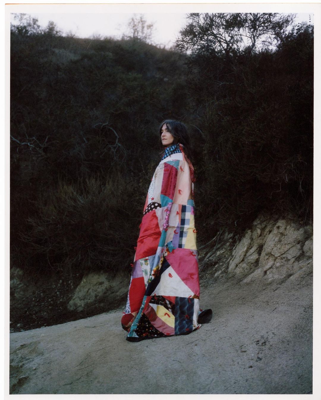 Woman in vibrant patchwork coat stands on a sandy path with a background of shrub-covered hills under a soft, overcast sky.