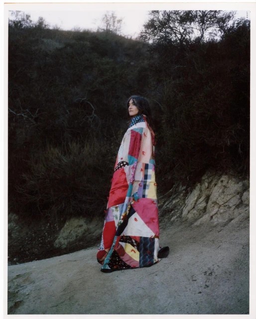 Woman in vibrant patchwork coat stands on a sandy path with a background of shrub-covered hills under a soft, overcast sky.