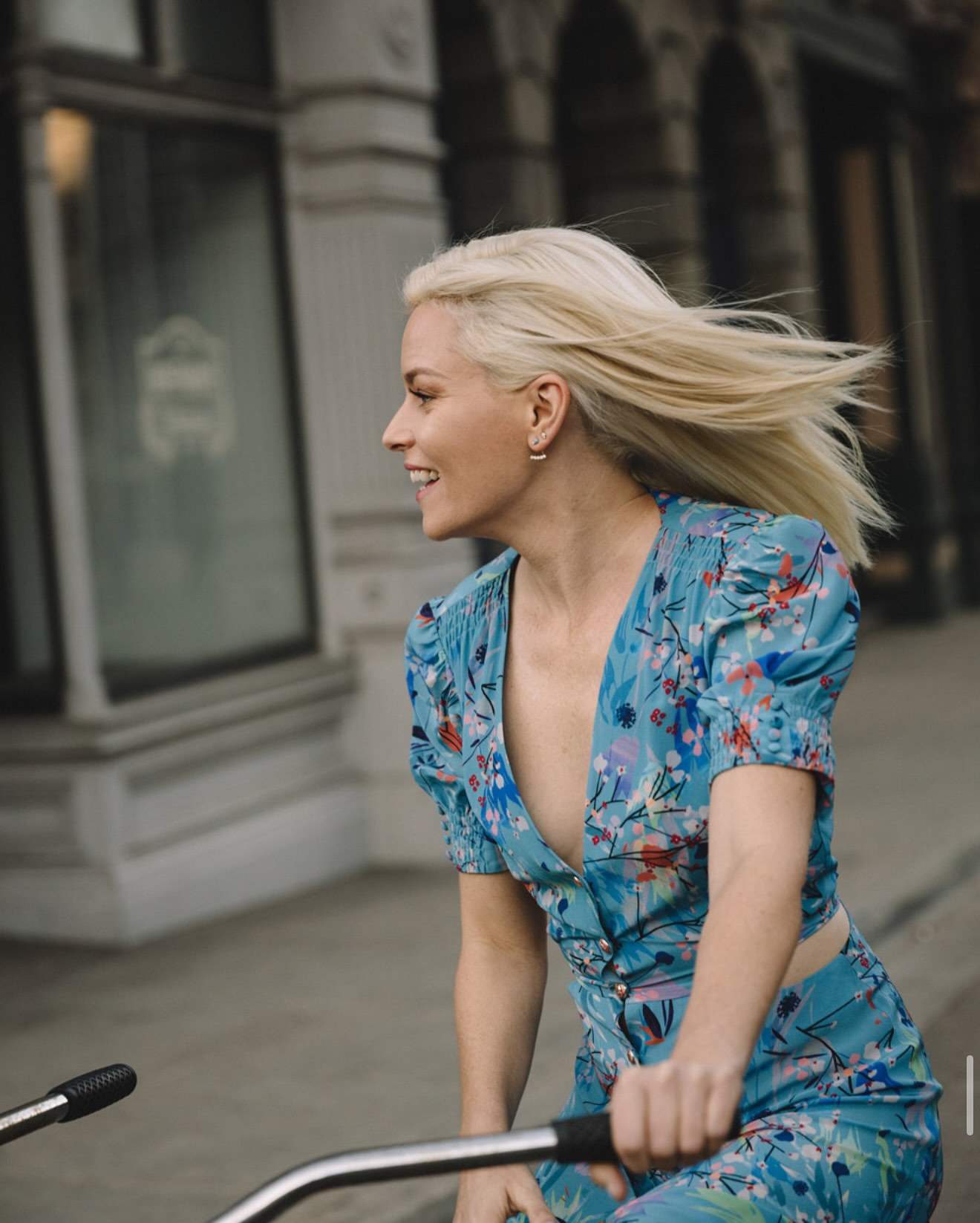 A woman with blond hair rides a bicycle outdoors. She wears a blue floral print outfit and smiles as she cycles in the city.