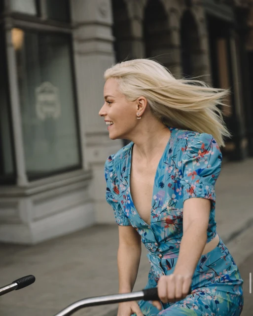 A woman with blond hair rides a bicycle outdoors. She wears a blue floral print outfit and smiles as she cycles in the city.