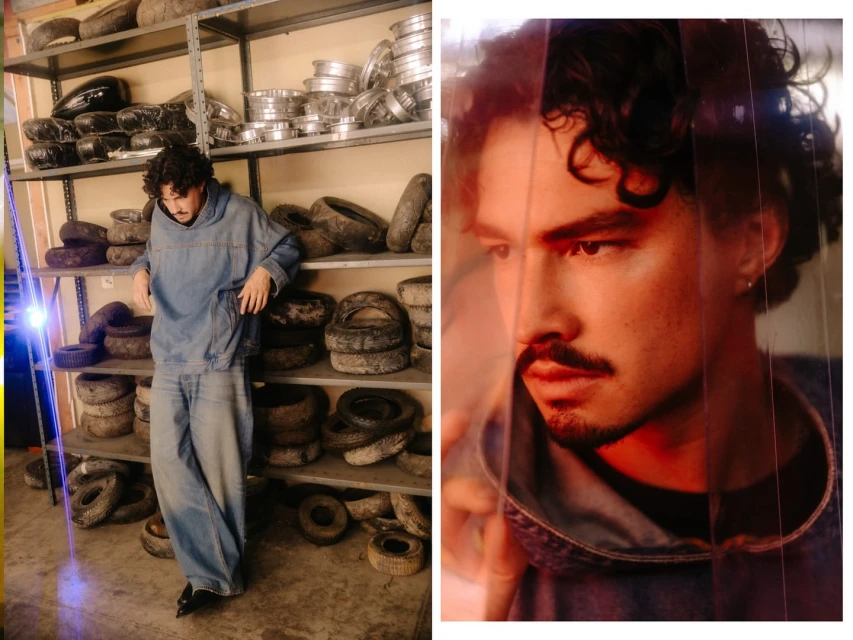 A stylish man in a denim outfit poses in a warehouse setting with tires and metal containers, captured in warm, natural light.