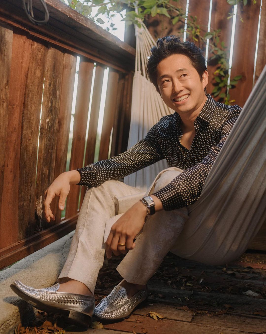 Portrait of Steven Yeun sitting in a hammock with a polka-dot button-down shirt, beige pants, and silver loafers outside.