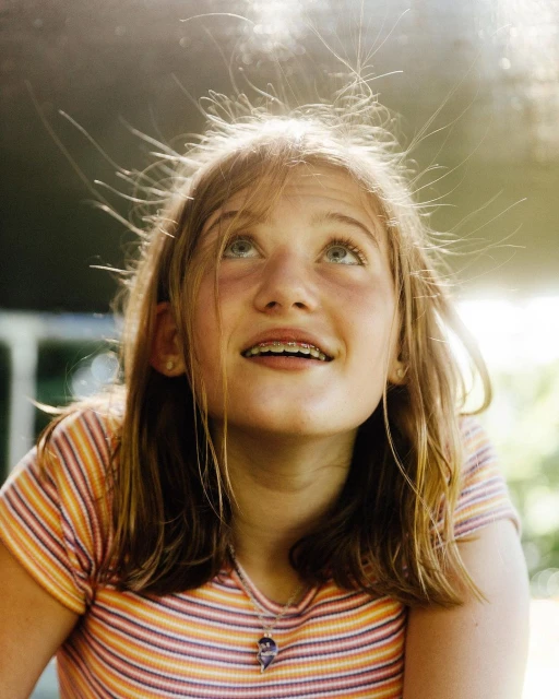 A smiling girl with braces looks upward, her hair standing on end from static electricity. Outdoor setting, striped shirt and pendant necklace.