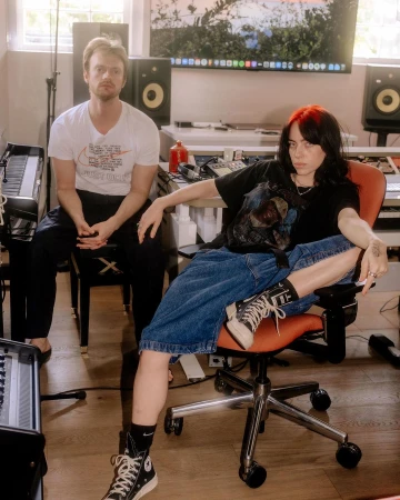 Two musicians, an adult male and female, are photographed in a music studio with musical instruments and equipment, posing together in casual attire.