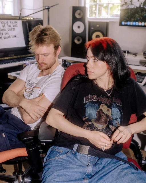Two musicians, a man and a woman with red-streaked hair, are seated in a music studio with speakers, piano, and handwritten lyrics in the background.