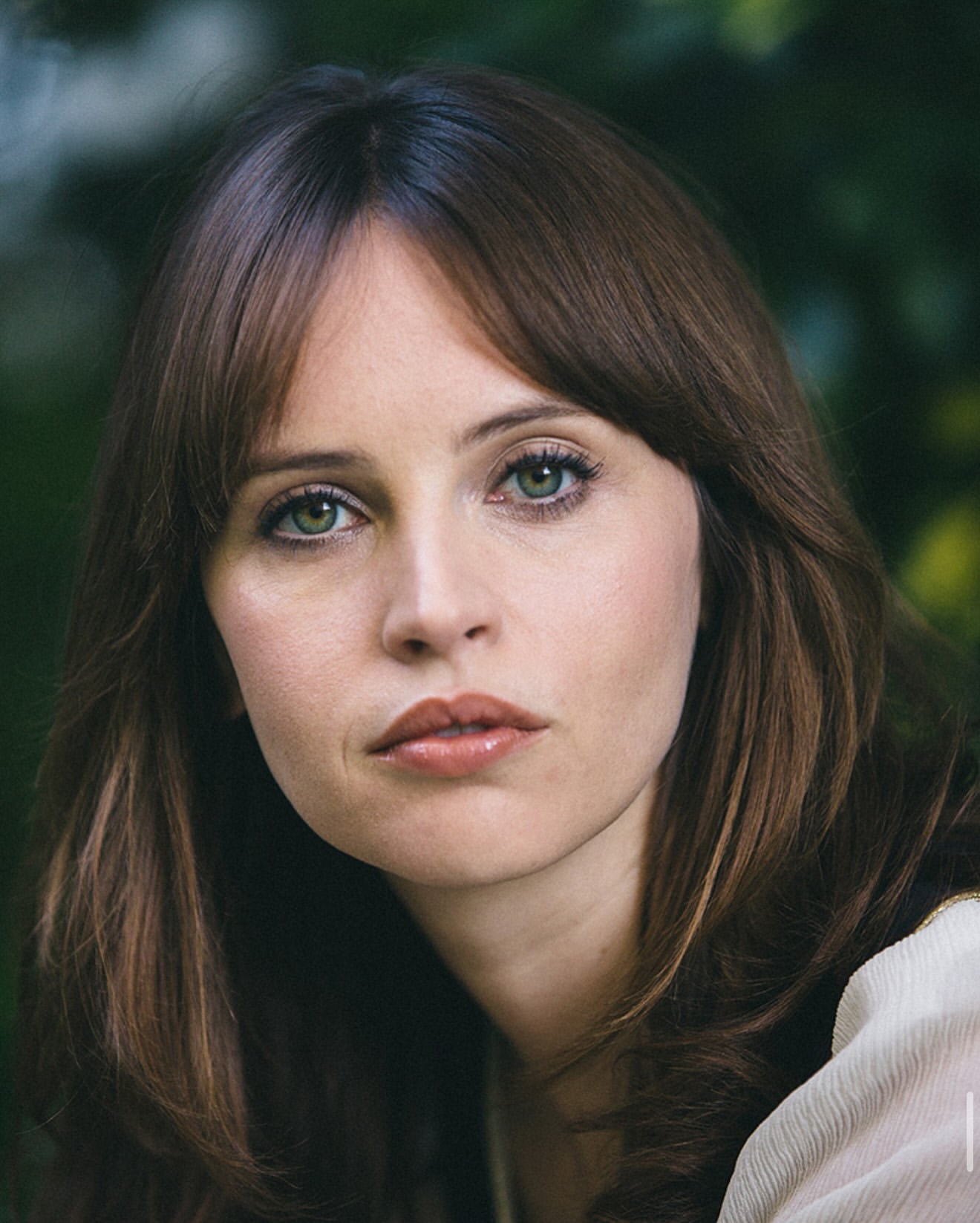 Portrait of Felicity Jones with brown hair, bangs, and green eyes outdoors with green foliage background.
