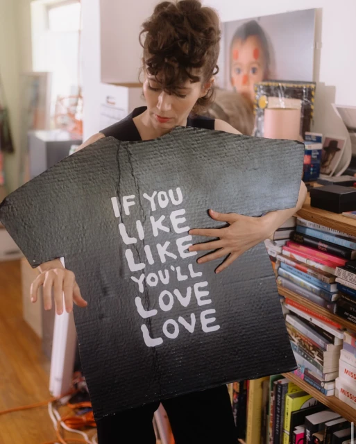 Adult woman holding a hand-painted sign saying, 'If you like like, you'll love love', standing indoors with books and shelves in the background.
