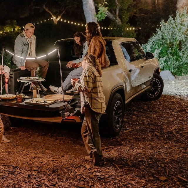 People gathered around a Rivian electric truck in an outdoor setting, illuminated by string lights at night, creating a social gathering.