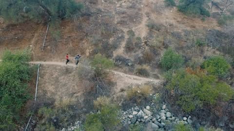 Two people trail running on a dirt path winding through a hilly landscape with trees and rocks on a sunny day.