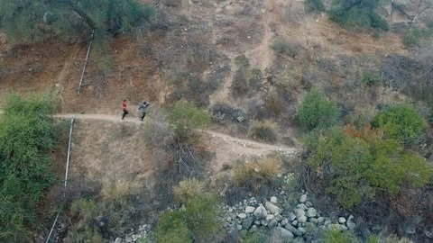 Two people trail running on a dirt path winding through a hilly landscape with trees and rocks on a sunny day.