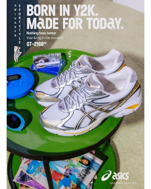 Two Asics GT-2160TM athletic shoes in white and silver are displayed with a CD player and cassette tapes on a two-tiered green table.
