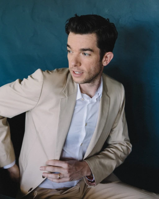 Adult male poses in a tan suit against a mottled blue backdrop in a studio shot. The subject gazes to the left.