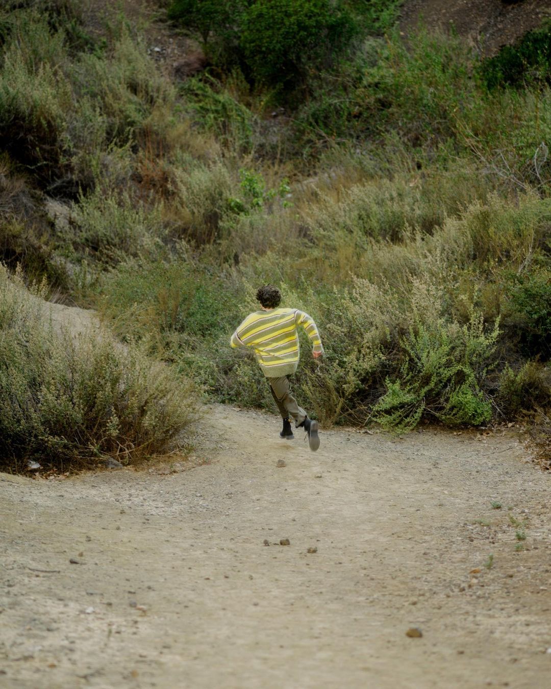 Person wearing a striped yellow and white sweater running down a dirt path through dense green bushes and overgrown terrain.