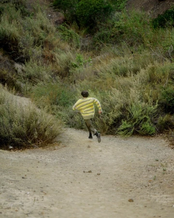 Person wearing a striped yellow and white sweater running down a dirt path through dense green bushes and overgrown terrain.