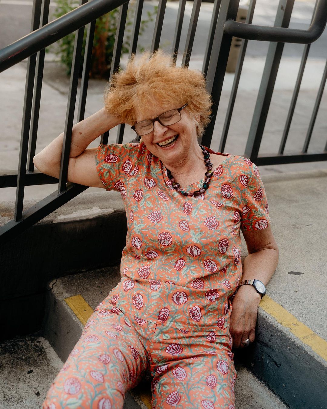 Elderly woman with short red hair smiles while sitting on concrete stairs, wearing a floral patterned top and pants and a beaded necklace.
