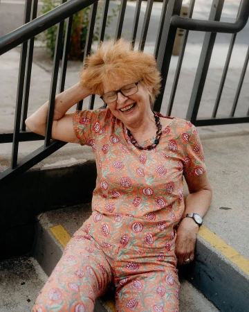 Elderly woman with short red hair smiles while sitting on concrete stairs, wearing a floral patterned top and pants and a beaded necklace.