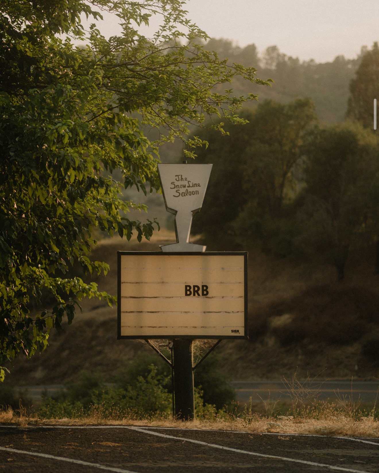 A sign for The Snow Line Saloon with the letters BRB on the bottom panel, surrounded by green trees and a road in the foreground.