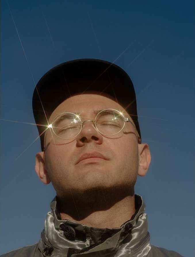 Portrait of an adult with closed eyes wearing gold-rimmed glasses and a black cap set against a clear blue sky background.