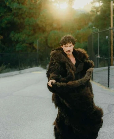 Man wearing a long dark faux fur coat standing on an asphalt street with a chain-link fence and warm sunlight in the background.