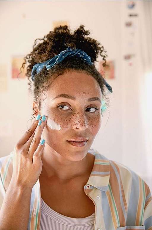 Woman applies lotion to her face indoors during the daytime. She has freckles, curly hair, and wears a polka-dot hair accessory.