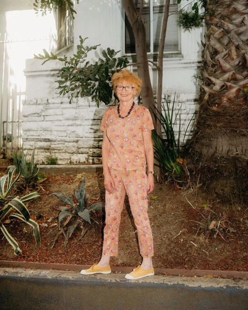 Full shot of a senior woman in a printed shirt and pants standing outdoors near a white building and greenery on a sunny day.