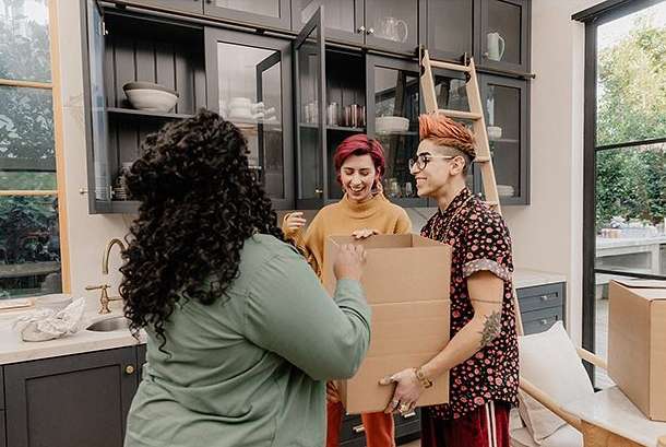 Three people unpack boxes in a kitchen, featuring two women and one man with a modern interior design and natural light from a window.