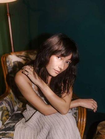Woman with long dark hair wearing a light-colored dress seated in an armchair against a dark green wall
