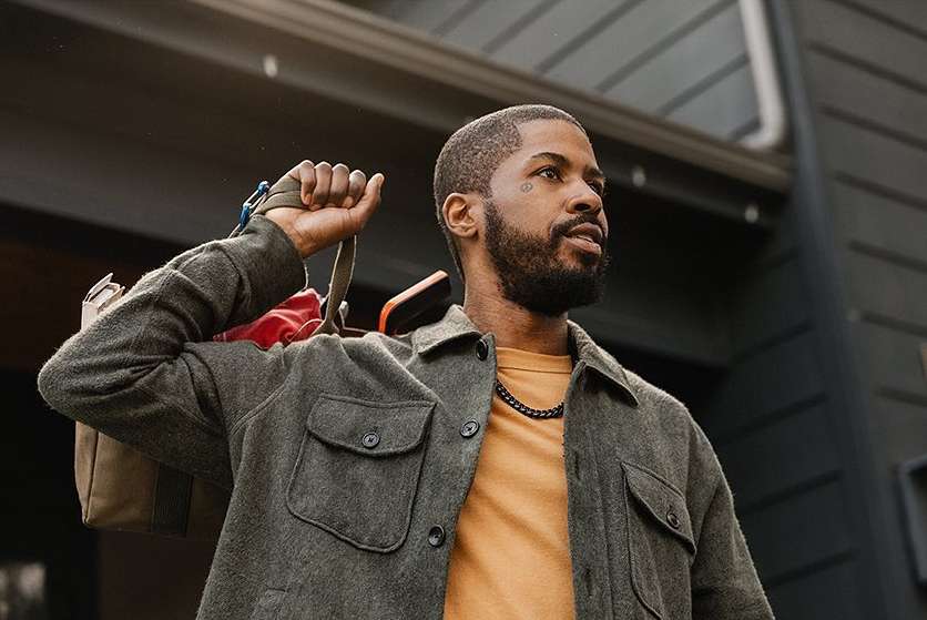 Stylish African-American man in casual gray jacket with bag over shoulder, standing outside gray house. Male fashion and lifestyle.