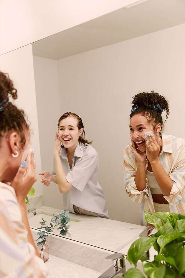 Two smiling adult women washing their faces with cleanser in a brightly lit bathroom with a white marble countertop and green plant.