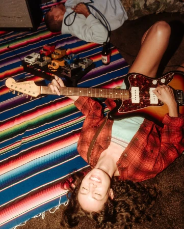 Young woman plays electric guitar while lying on a striped blanket, with a man resting nearby, creating a relaxed musical scene.