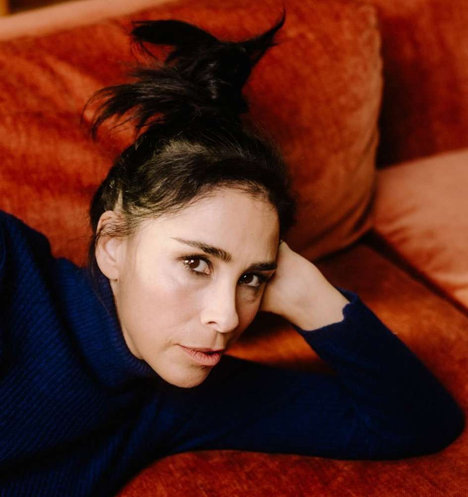 Woman with black hair reclines on an orange couch, wearing a dark blue sweater, captured in a close-up, indoor setting.