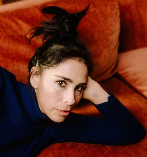 Woman with black hair reclines on an orange couch, wearing a dark blue sweater, captured in a close-up, indoor setting.